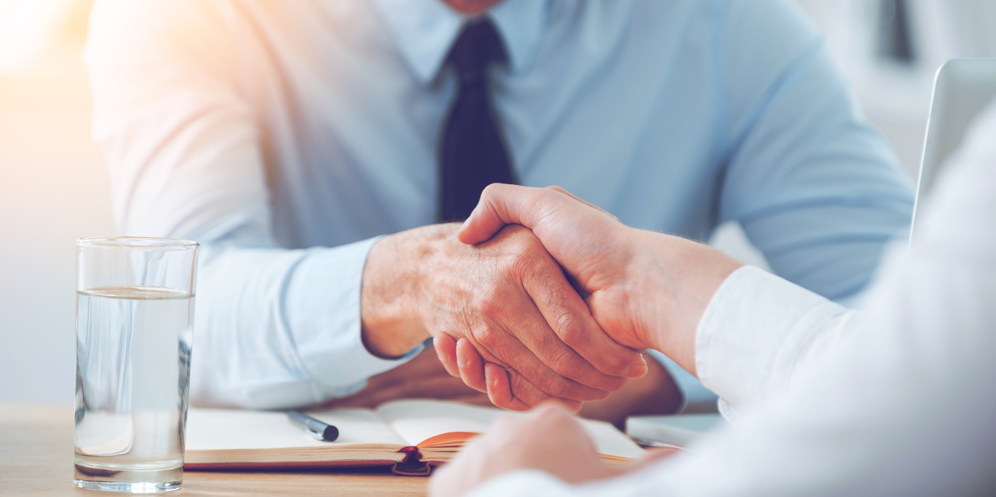 Two individuals shake hands over a table, symbolizing a business agreement. A glass of water and an open notebook are visible, indicating a professional setting.