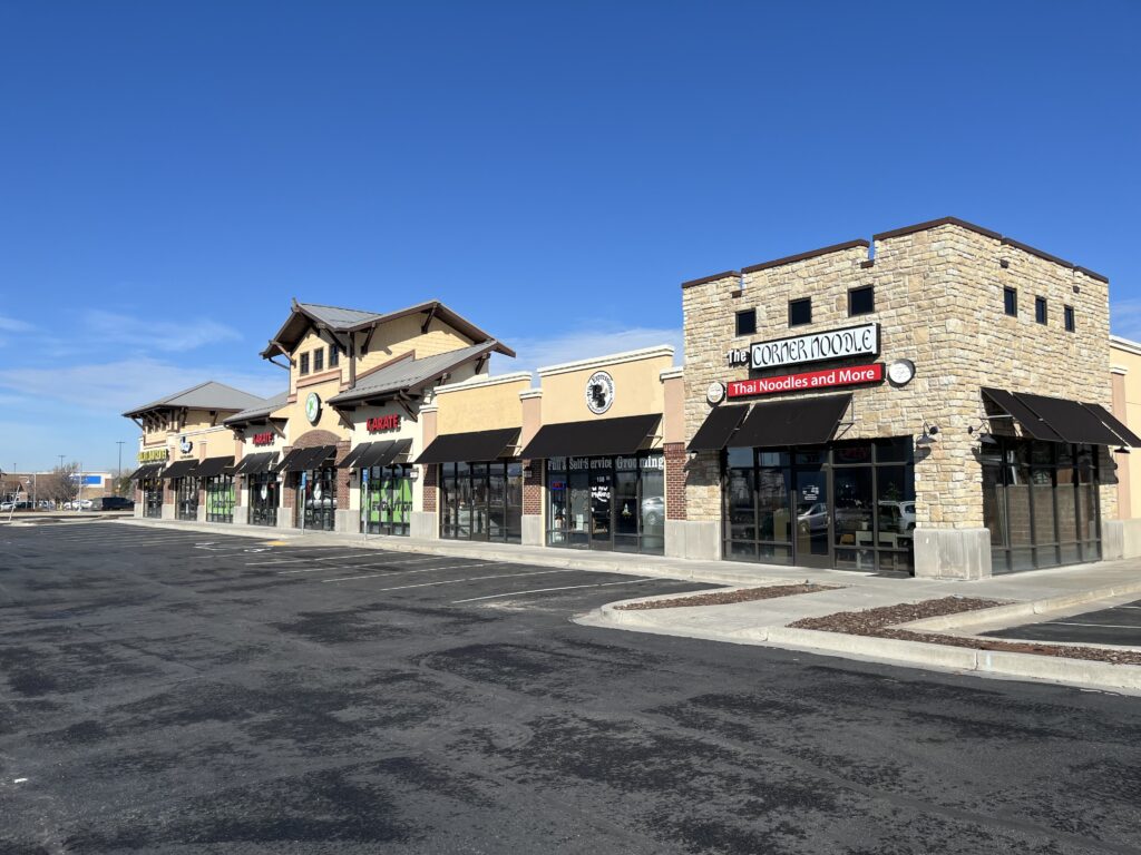 Row of businesses featuring The Corner Noodle restaurant with Thai cuisine. Clear blue sky and empty parking lot suggest a quiet atmosphere.