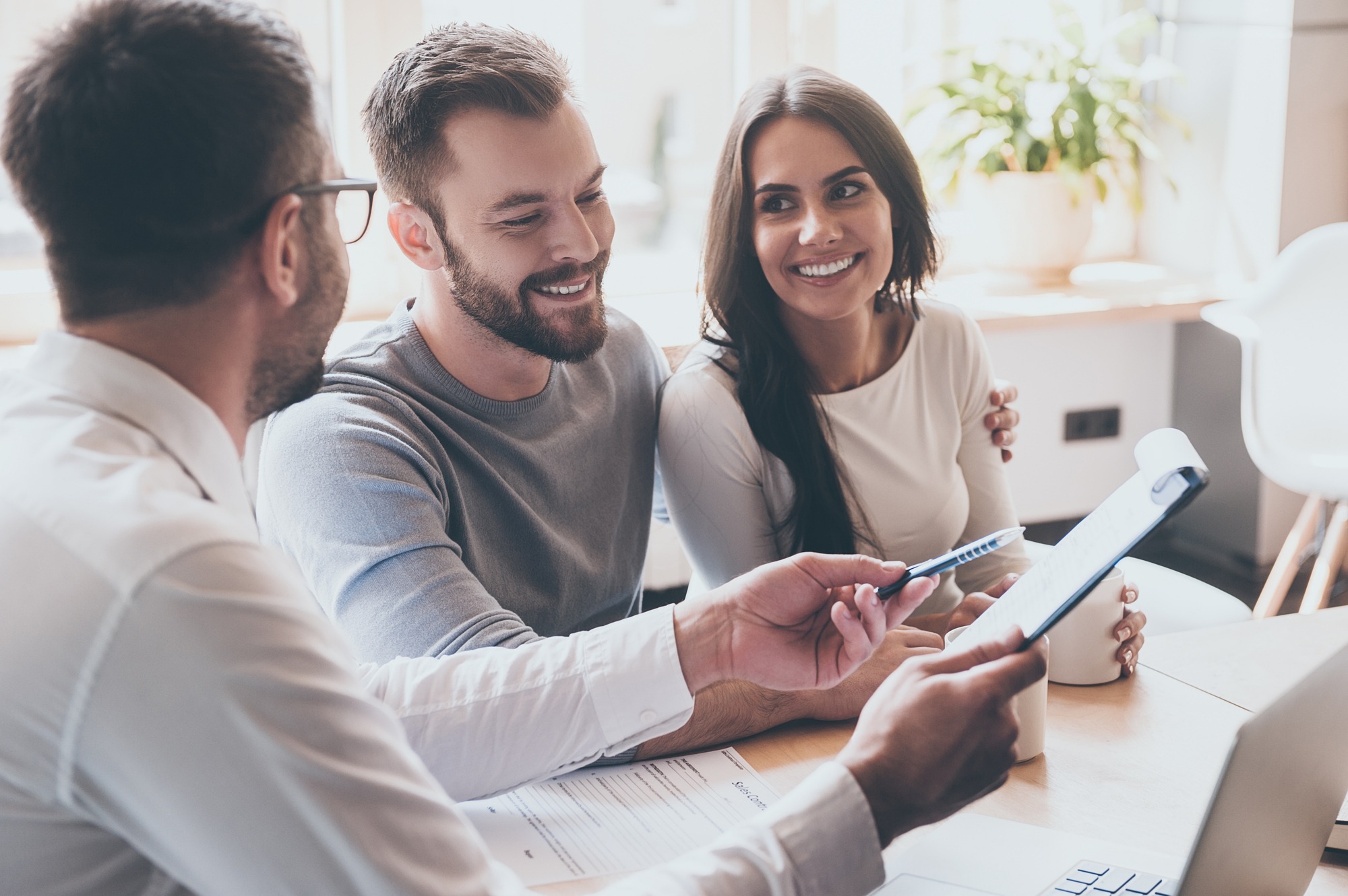 Three people are engaging in a friendly discussion around a table, reviewing documents. The atmosphere is collaborative and professional.